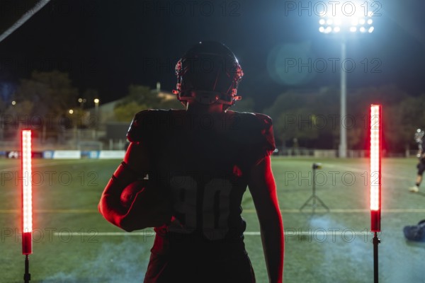 Football player stands on stadium field at night, helmet and uniform on, holding ball and facing bright red glowing lights, poised and ready for intense competition and victory