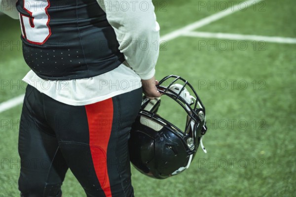 American football player standing on a green turf field, holding a black helmet and wearing a uniform with jersey and pants, ready for competition or reflecting on the game