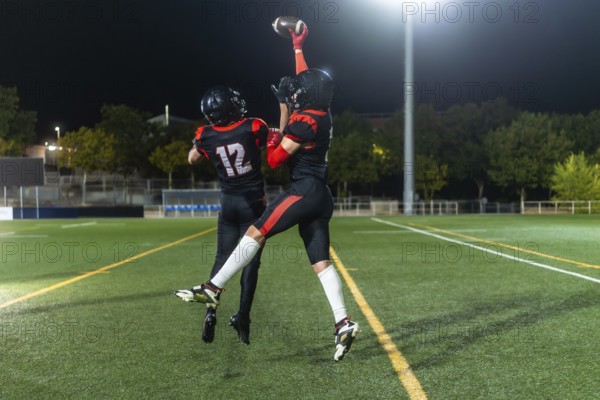 American football players wearing helmets and jerseys leaping to catch a football during a night game on a floodlit stadium field, showing intense action and competition