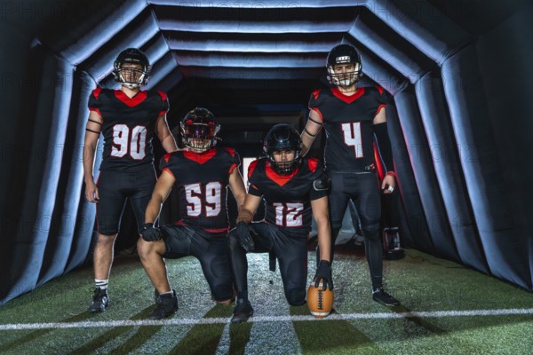 American football players in black and red uniforms, some kneeling and some standing in a dark tunnel holding a ball, poised to charge onto the field with focused determination