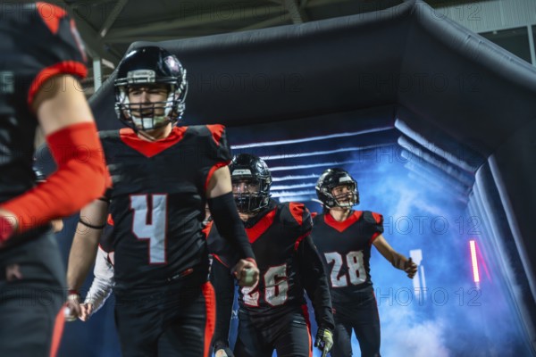 American football players, wearing black and red uniforms with helmets, walking into the stadium field through an inflatable tunnel filled with blue and pink smoke, ready for the game