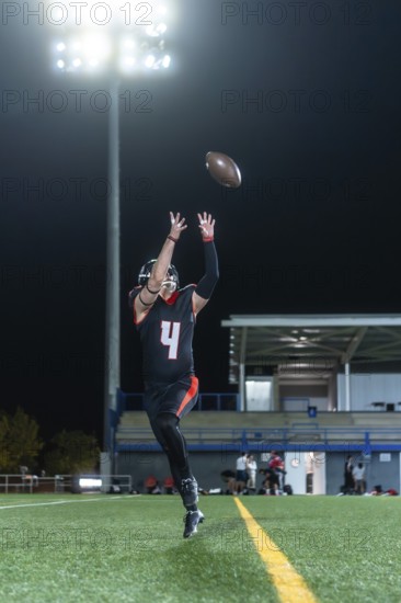 American football athlete in uniform and helmet jumping during a night game on a stadium field, attempting to catch an airborne ball under bright lights