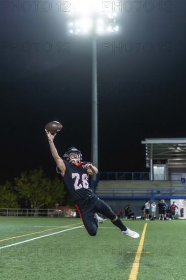 American football player in uniform and helmet is mid air, making a dynamic catch of a pigskin ball while playing on a green stadium field at night under bright overhead lights