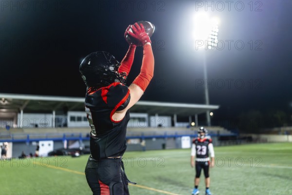 American football player in red and black uniform and helmet stretches arms to catch ball under floodlights during a nighttime stadium game, showing focus, power and motion