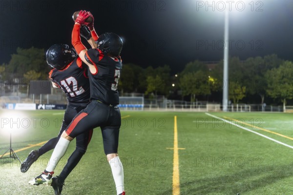 Two female american football players in black uniforms leap to catch the ball during a night game under stadium lights, showcasing speed, power, teamwork and determination