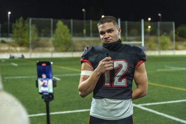 American football player in uniform number 12, standing on a green sports field at night, holding a lavalier microphone while recording an interview on a smartphone set up on a tripod