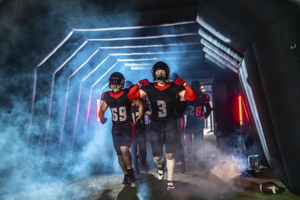 American football players in black and red uniforms and helmets running through a dramatic, smoke filled stadium tunnel with glowing lights, preparing for a game