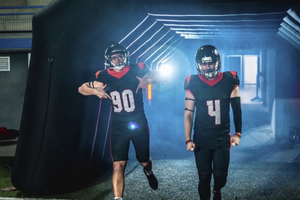 American football players in black and red uniforms and helmets are making an entrance, emerging from a dark tunnel illuminated with blue light, ready for a game on the field below