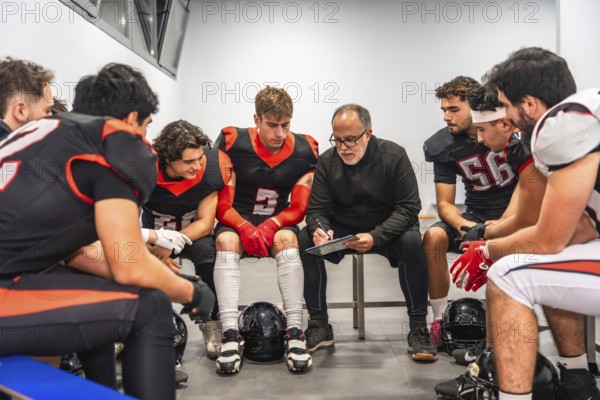 American football coach discussing strategy with his team of male players in the changing room, analyzing tactics and giving instructions before a game