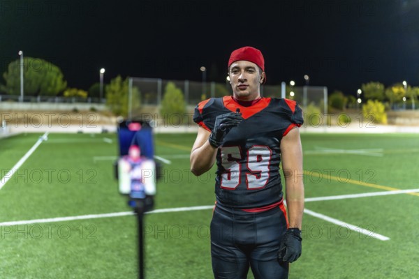 Football player in uniform recording video on a smartphone with a lavalier microphone, creating new content for social media and online channels at night on an artificial turf field