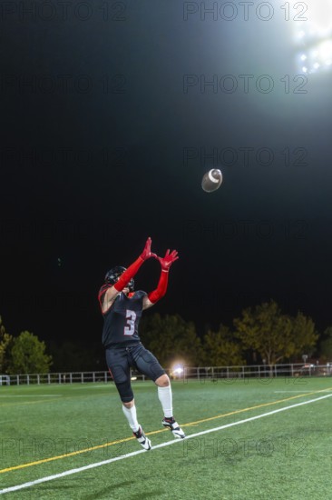 American football player in uniform and helmet is jumping to catch a football during a night game on a sports field under bright stadium lights, demonstrating athletic skill
