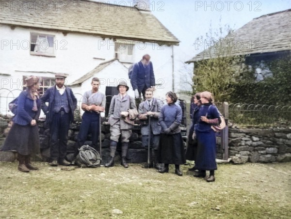 Colourised image of men and women in mountaineering mountain climbing group standing in fron of cottage, c 1900-1920