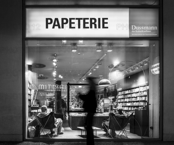 Black and white photograph, Dussmann GmbH, bookshop, urban scene in the evening with reflections in the window pane and a view of the displays and shelves, Berlin, Germany