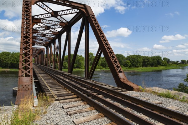 Railroad tracks leading to old rusted Terrebonne Pratt truss railroad bridge over Des Mille-Iles river in summer, Laval, Quebec, Canada