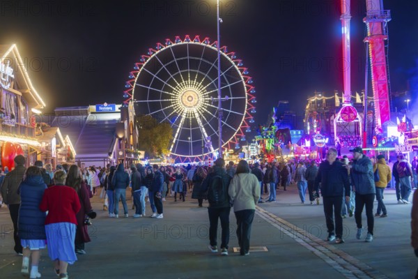 People walking on a festive square with a glowing Ferris wheel, Cannstatter Wasen folk festival, Stuttgart, Germany