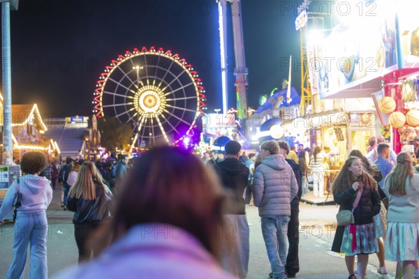 View of a busy fairground at night with an illuminated Ferris wheel and people waiting to go for a walk, Cannstatter Wasen fair, Stuttgart, Germany