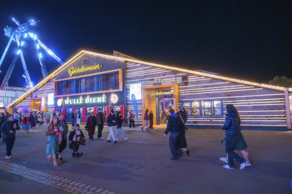 Visitors walk past an illuminated building at the funfair at night, Cannstatter Wasen funfair, Stuttgart, Germany