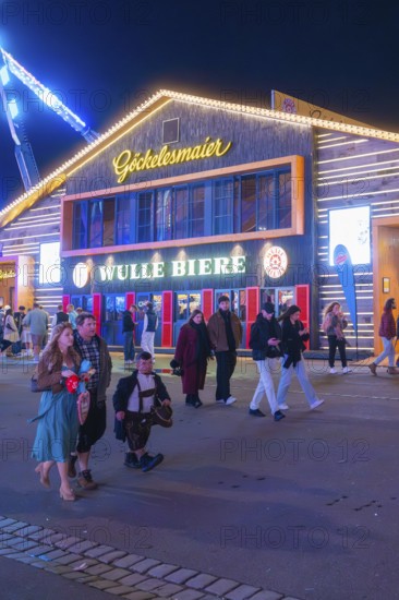 People strolling at night in front of an illuminated building at the funfair, Cannstatter Wasen folk festival, Stuttgart, Germany