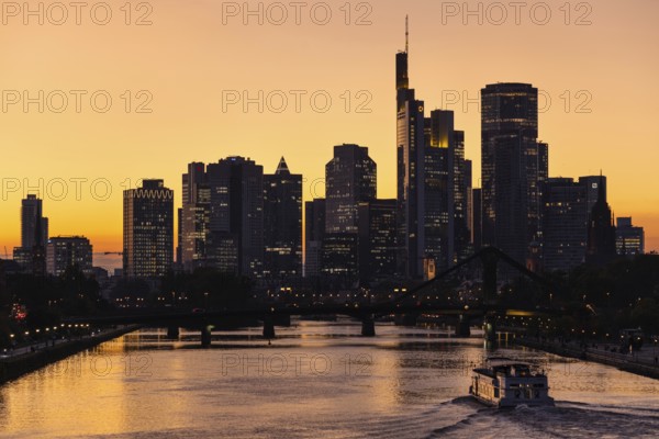 As a ship travels along the Main, the evening sky behind the Frankfurt banking skyline glows orange and yellow shortly after sunset, Frankfurt am Main, Hesse, Germany