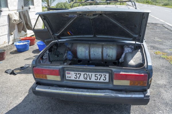 Old car with open boot, gas bottle and tools on a sunny day next to the road, car has gas tank retrofitted to be able to drive more cheaply, Armenia