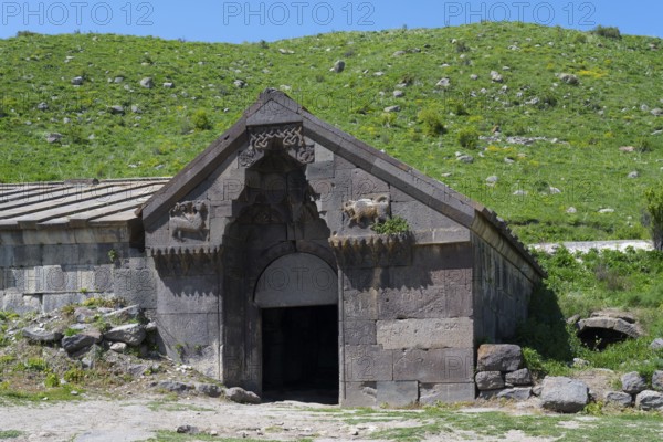 Old stone building with detailed decorations in front of a lush green hill on a sunny day, Orbelian's Caravanserai, Selim Caravanserai, located at Vardenyats Pass or Selim Pass, Vayots Dzor Province, Armenia