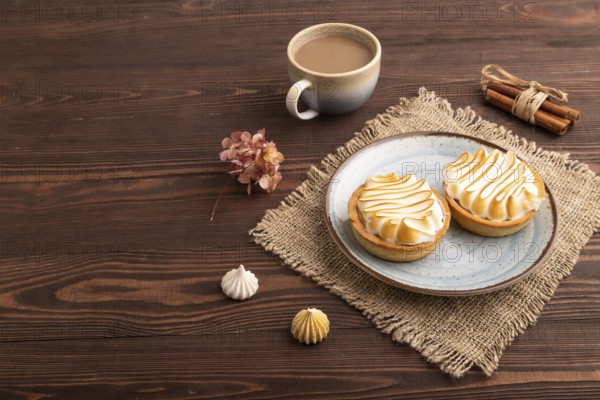 French lemon tart with meringue on brown wooden background, cup of coffee, linen textile, side view, copy space