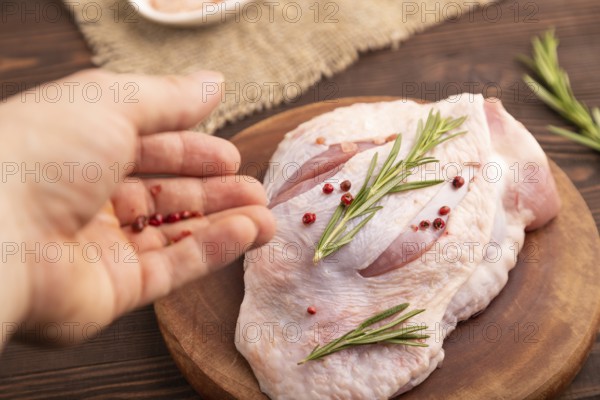 Raw Turkey Thigh with spices and rosemary on cutting board with hand on brown wooden background and linen textile. side view, close up, selective focus