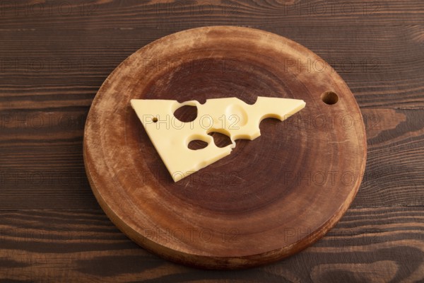 Piece of cheese with holes on cutting board on brown wooden background, side view, close up, minimalism