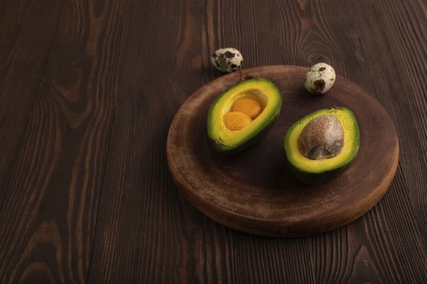 Two Avocado halves with pit and Egg yolk on round wooden cutting board on brown wooden background, side view, copy space, minimalism, low key
