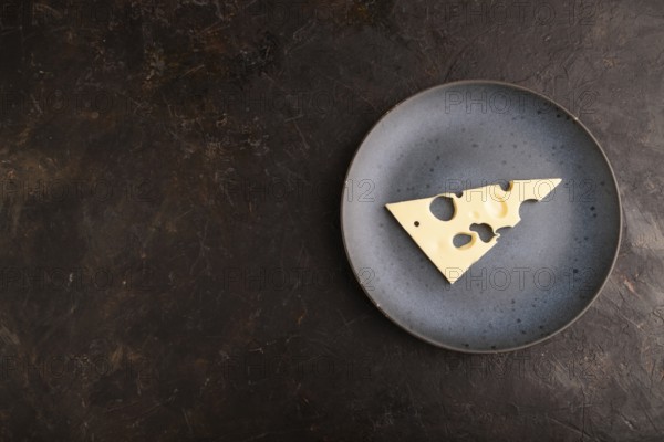 Piece of cheese with holes on blue plate on black concrete background, top view, flat lay, copy space, minimalism