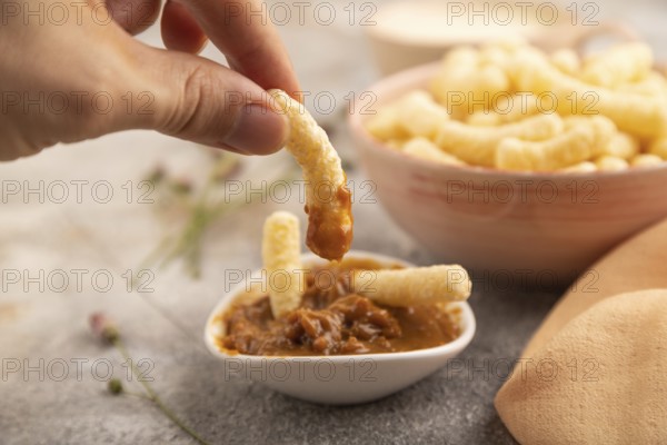Corn flakes sticks with hand with caramel in ceramic bowl on gray concrete background and orange linen textile. Side view, close up
