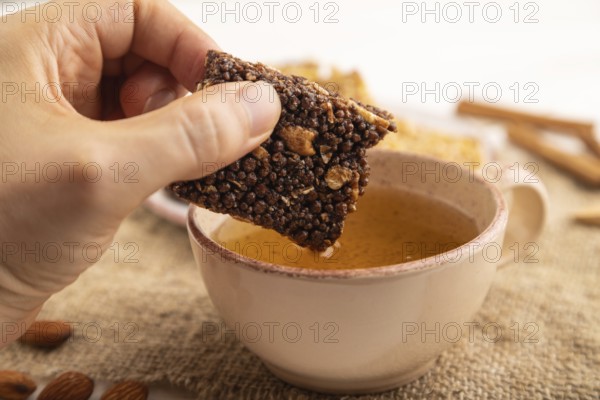 Granola bars with caramel, nuts, flakes in ceramic plate with hand on white wooden background, beige linen napkin, cup of green tea. Side view, close up, selective focus