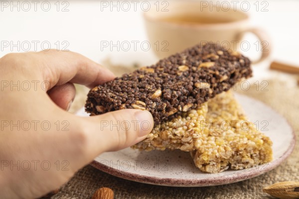 Granola bars with caramel, nuts, flakes in ceramic plate with hand on white wooden background, beige linen napkin, cup of green tea. Side view, close up, selective focus
