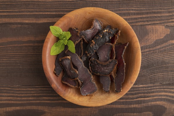 Armenian Basturma dried meat on wooden bowl with pepper and herbs on brown wooden background. Top view, flat lay, copy space
