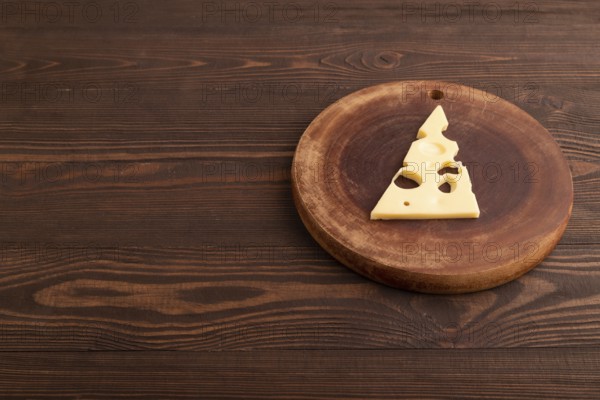 Piece of cheese with holes on cutting board on brown wooden background, side view, copy space, minimalism
