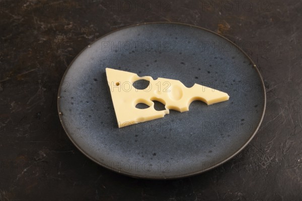 Piece of cheese with holes on blue plate on black concrete background, side view, close up, minimalism