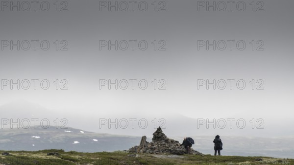 Two pilgrims lay stones at the stone pyramid Allmannrøysa, cloudy landscape, pilgrimage route Olavsweg or Olavsleden, old Kongevegen or King's Path over the Dovrefjell, Norway