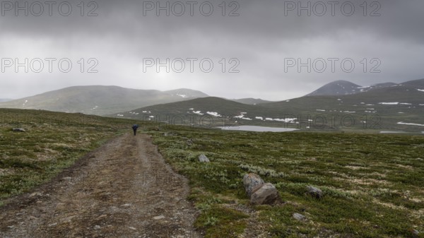 Hiker with rucksack and blue raincoat on a lonely plateau with snowfields, low clouds, Kongevegen or King's Road, Olav's Way or Olavsleden, Dovrefjell, Oppdal municipality, Trøndelag county, Norway