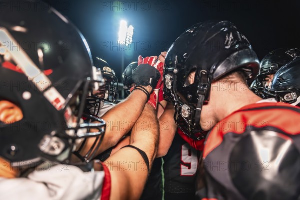 American football players are huddling on the field under stadium lights at night, putting their hands together in a gesture of unity, strategy, and team spirit before a game