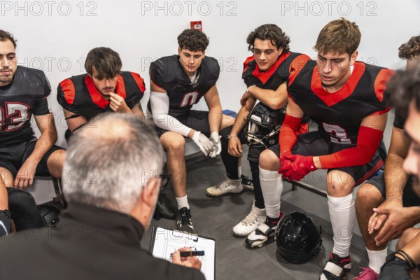 Football coach reviews clipboard plays while focused young players sit in the locker room, listening intently as he gives tactical instruction and motivation before the game