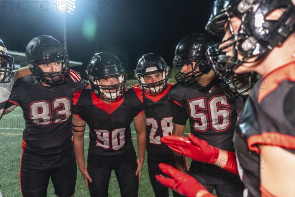 American football players huddling on a lit turf field at night, helmets on, focused on strategy and teamwork as coach and teammates prepare for the next play