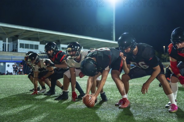 American football players in full uniform and helmets lining up on the green grass field at night, preparing for the game to start under bright stadium lights