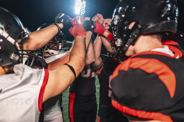 American football players huddling on the illuminated field, placing their hands together in the center, demonstrating teamwork, unity, and determination during a night game