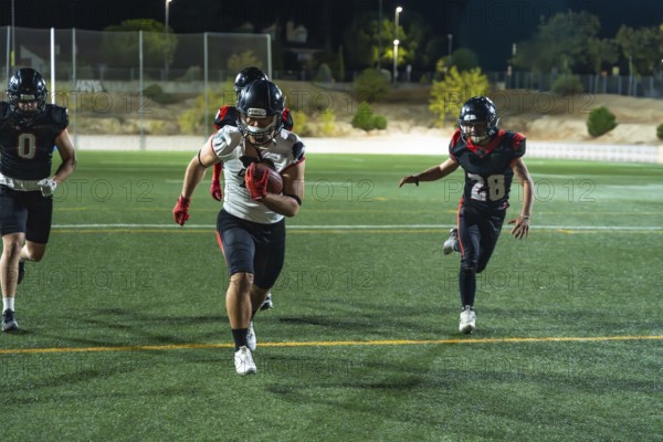 American football players competing on a lit field at night, a player carrying the ball and sprinting to gain yards while opponents are pursuing him during intense action