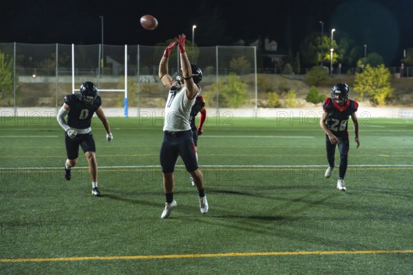 American football players are competing on a green field at night, a receiver is jumping mid air with outstretched arms attempting to catch a thrown football during a game