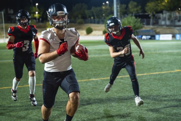 American football players, wearing protective helmets and pads, are running on a green turf field under bright stadium lights during an intense training session or evening game