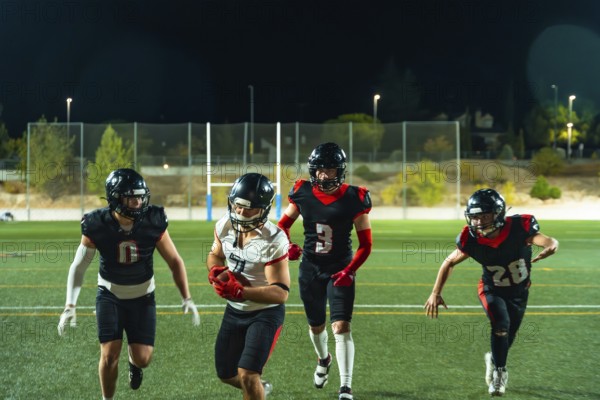 Four young american football players are running on the field under stadium lights at night, with one player carrying the football, showing teamwork and active play