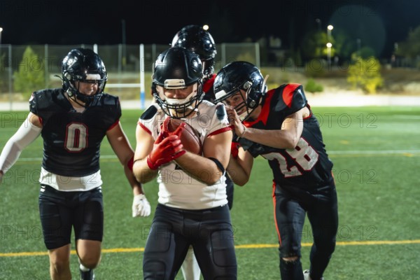 American football players in uniform and helmets engaging in a game on an illuminated sports field at night, one player holding the ball while others compete for it