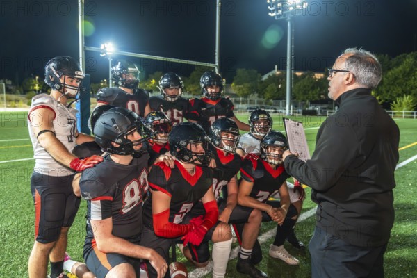 American football coach reviewing game strategy and tactical plays with a kneeling team of young players on a field illuminated by stadium lights at night