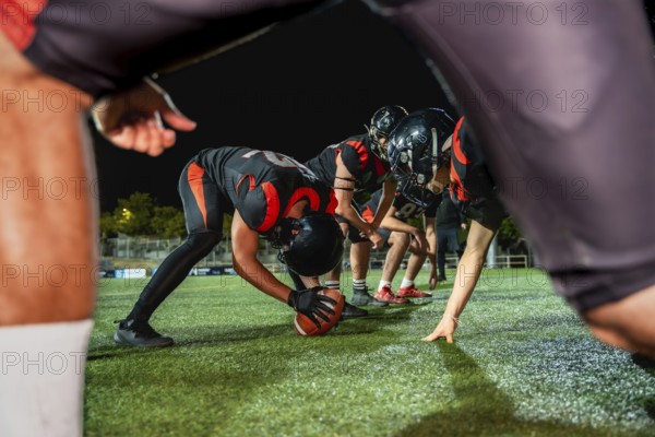 American football players line up on the field at night, a center snapping the ball, preparing for an intense moment of competitive sports action during a game